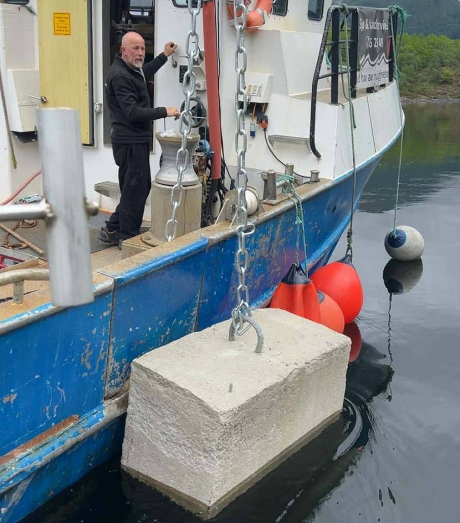 En ilestein i betong henger i kjettingen langs styrbord side på M/S Plutselig, klar til å senkes. En mann betjener vinsjen på dekk.